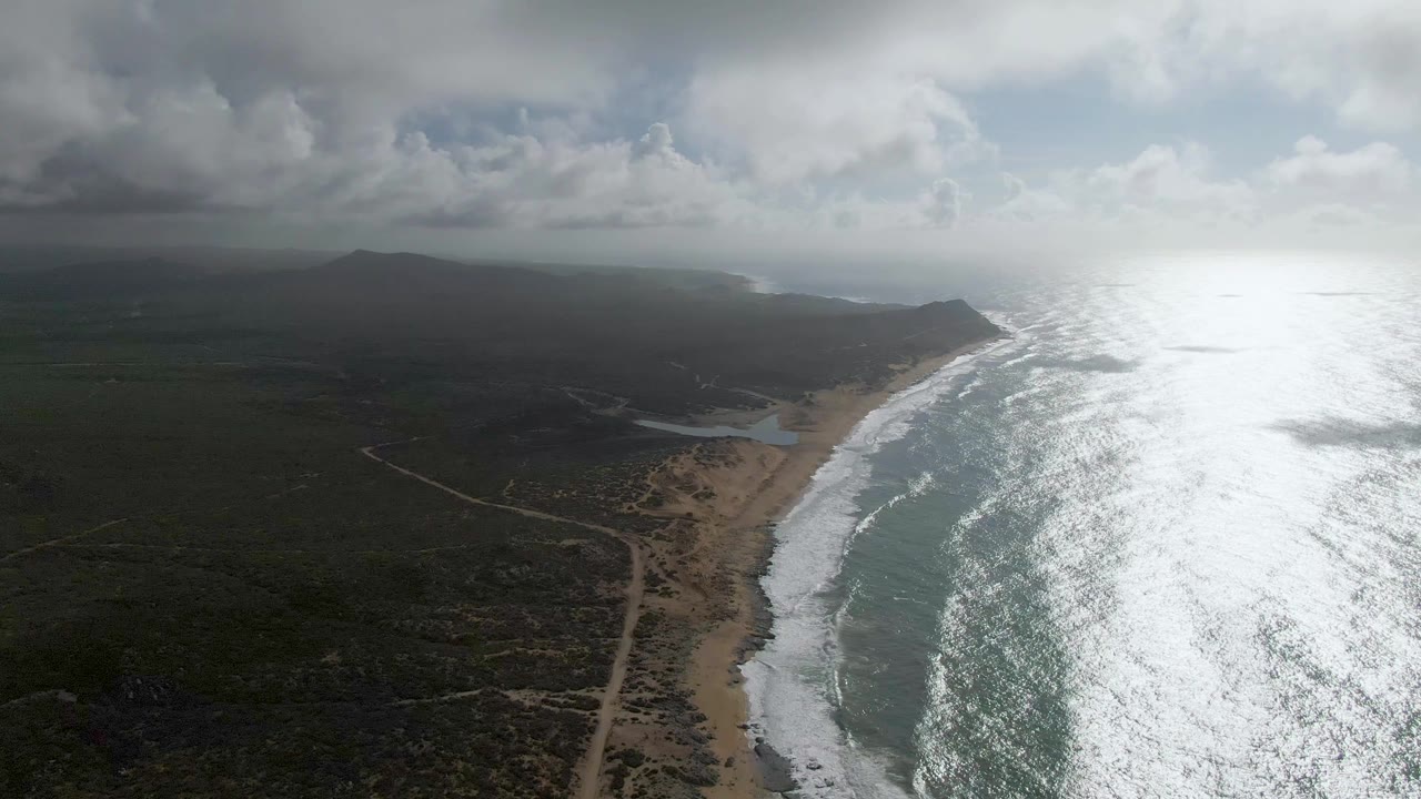 Aerial view of Punta Gorda in Baja California Sur showing bright surf on a sandy shoreline, a small lagoon near the beach, dark inland scrub, distant mountains, cloudy sky, and no visible people.