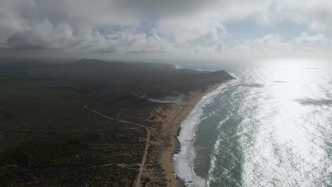 Aerial view along the Punta Gorda shoreline with reflective ocean occupying the right side, white surf tracing the beach, dark vegetation inland, distant mountains under heavy clouds, and no visible people.