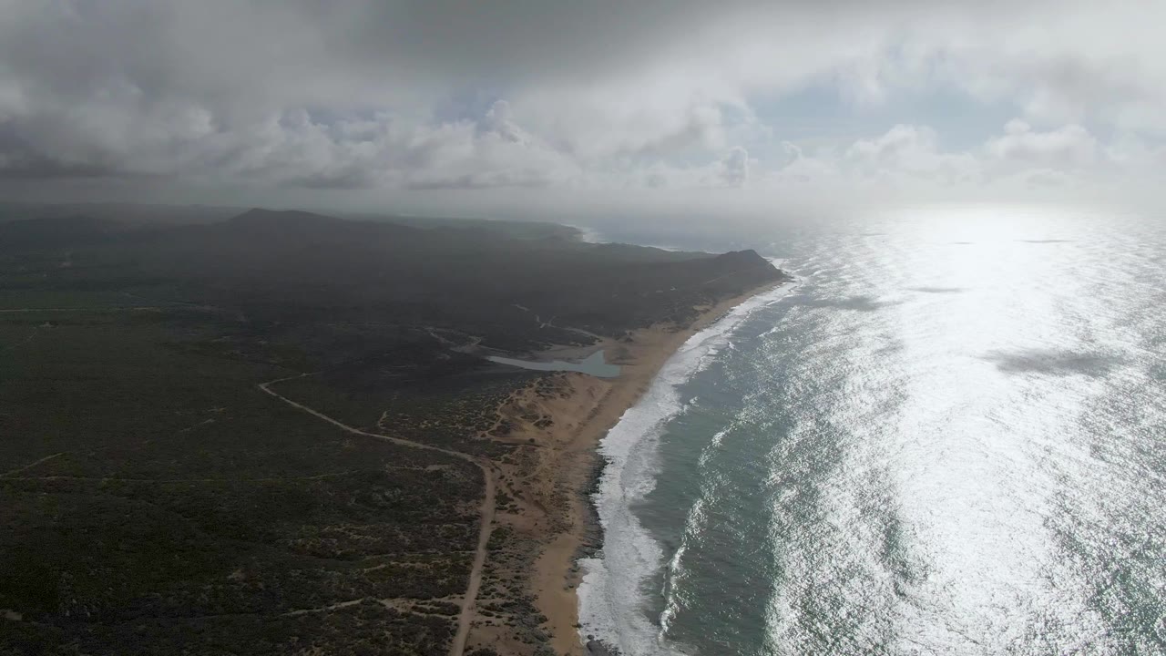 Aerial coastline with sandy beach, surf, dark land, clouds, and bright ocean reflection.