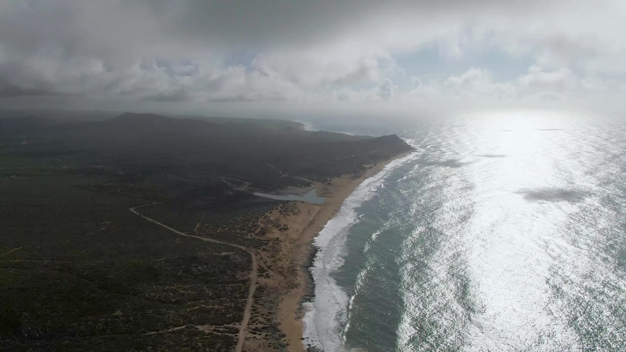 Aerial frame at Punta Gorda with the diagonal beach line, white wave bands, inland dirt roads, cloud-darkened vegetation, bright sea glare, and no visible people.