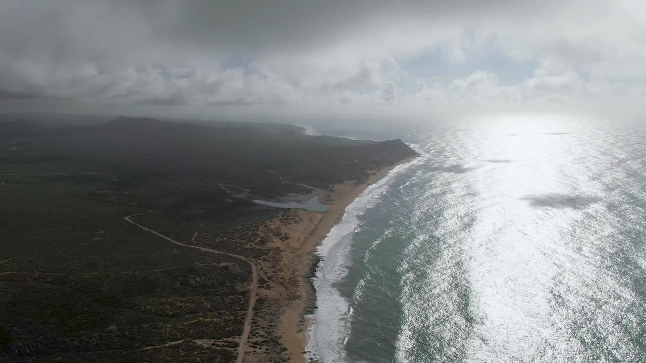 High drone perspective over Punta Gorda with a curved sandy beach, rolling white surf, broad reflective ocean, muted inland terrain, low clouds, and no visible people.