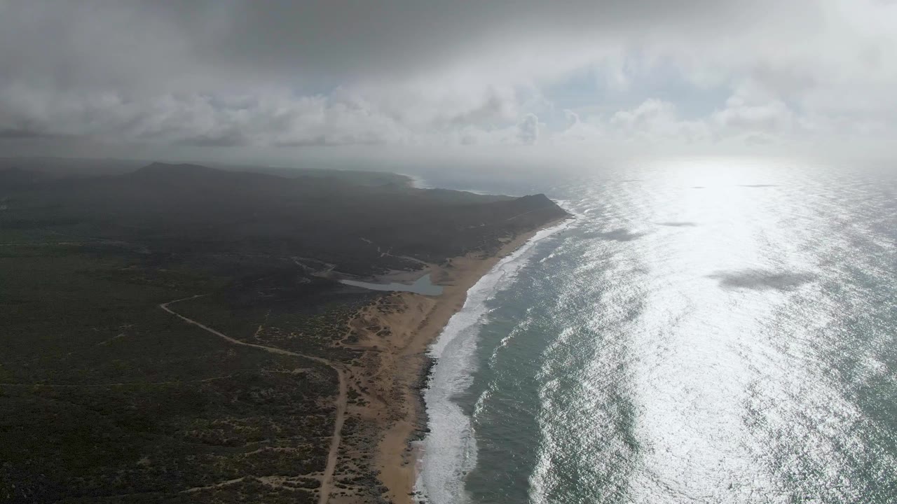 Punta Gorda aerial view with dark clouds across the upper frame, a sandy beach below, white surf along the shore, faint dirt roads inland, bright ocean reflection, and no visible people.