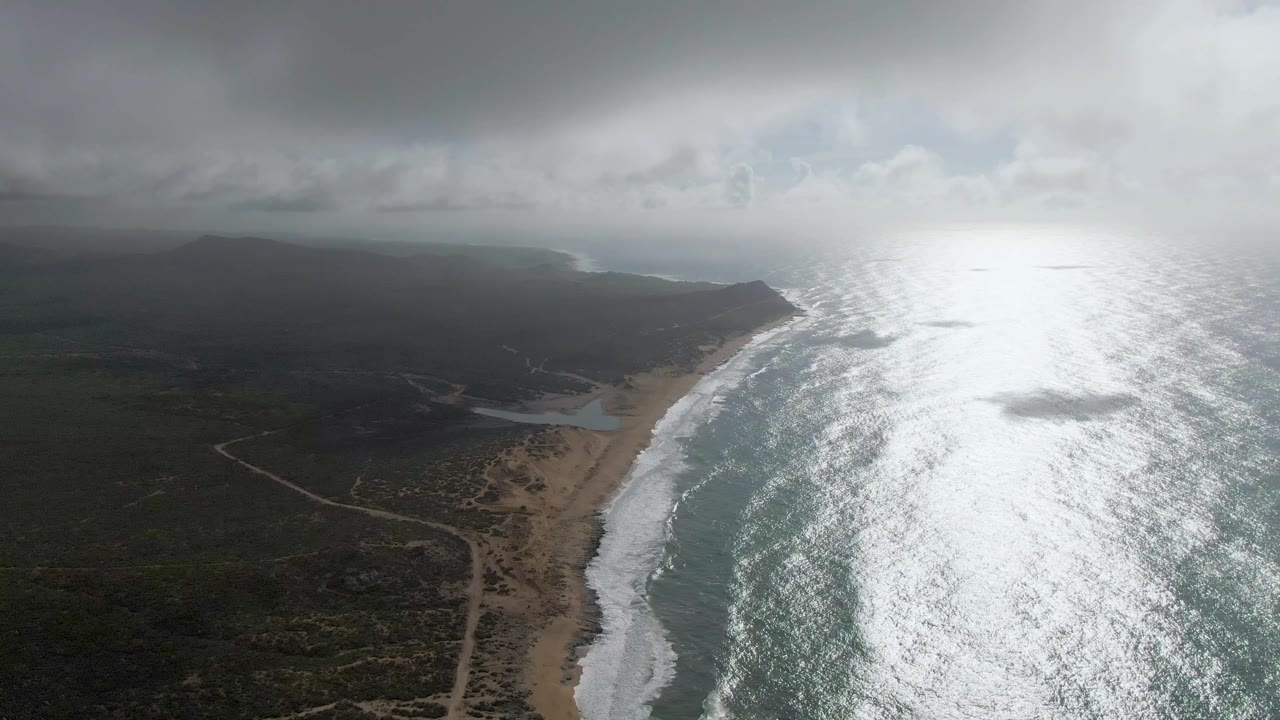 Aerial view of a sandy coastline, surf, scrubland, and bright ocean glare under clouds.