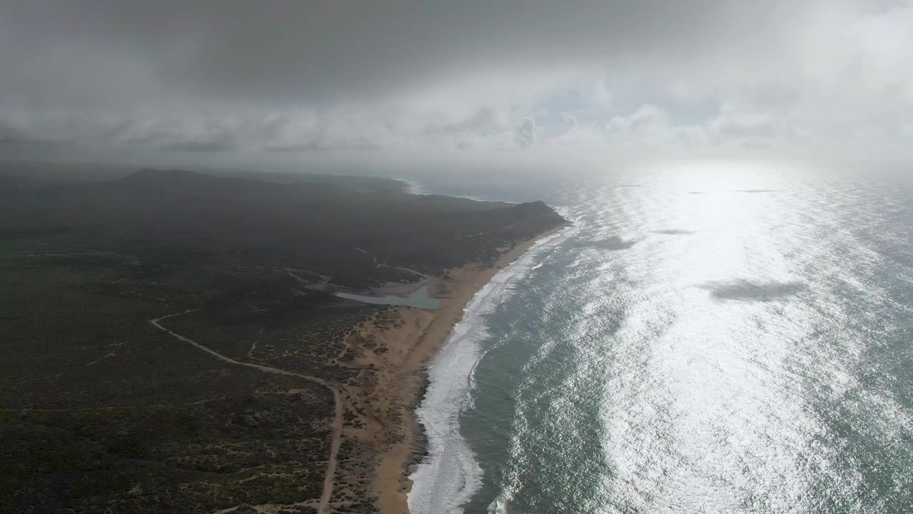 Aerial coastal frame from Punta Gorda, East Cape, showing the beach curve and breaking waves beside dark scrubland, with the ocean reflection shifted slightly across the right side and no visible people.