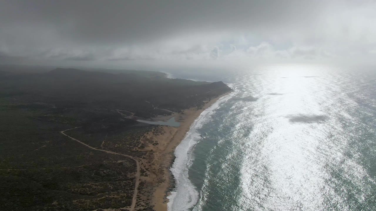 Aerial view over Punta Gorda, East Cape, with pale dirt roads crossing coastal scrub toward the beach, waves breaking along the sand, strong ocean glare to the right, distant dark land, and no visible people.