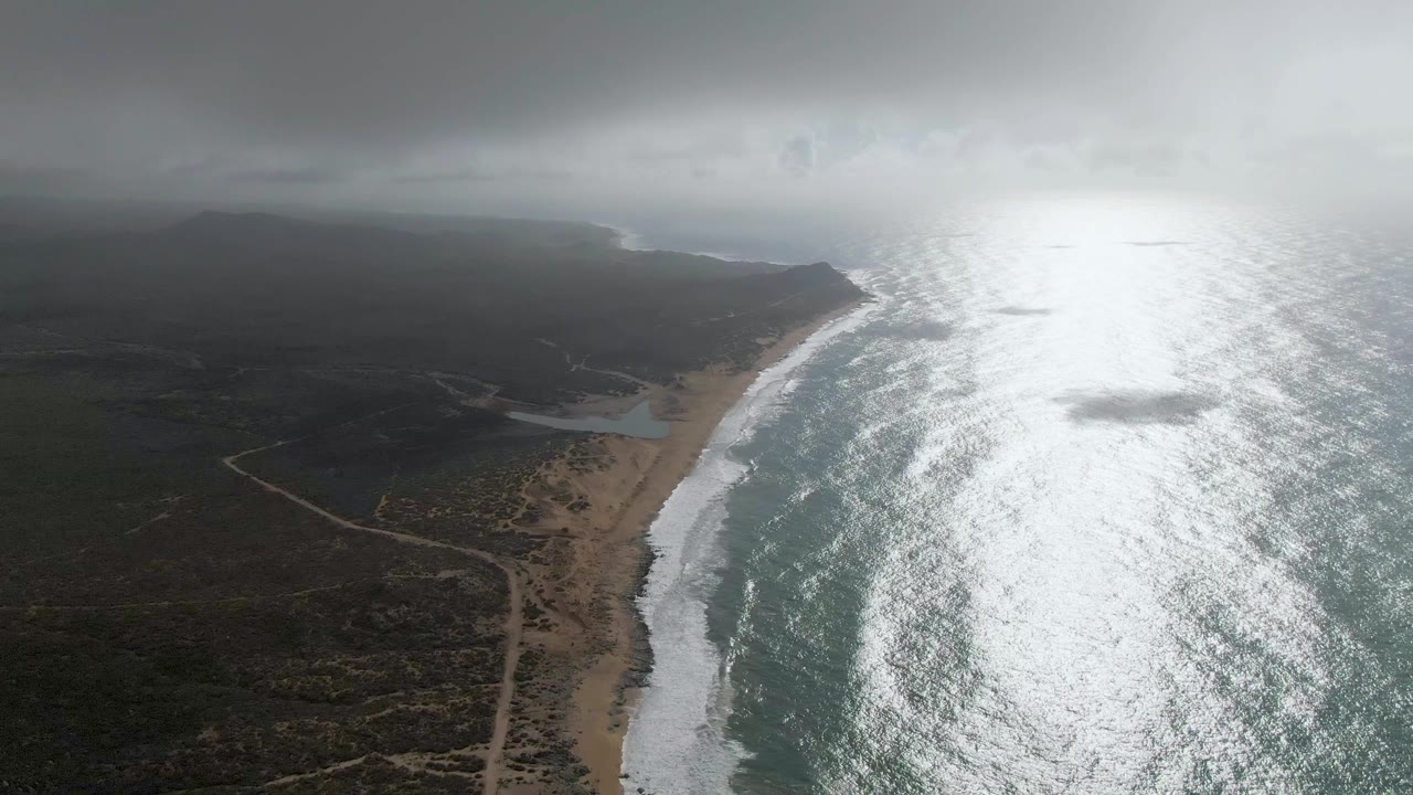 Aerial view of Punta Gorda East Cape coastline with sandy beach, white surf, scrub-covered land, dirt roads, bright ocean reflection, low cloud, and no visible people.