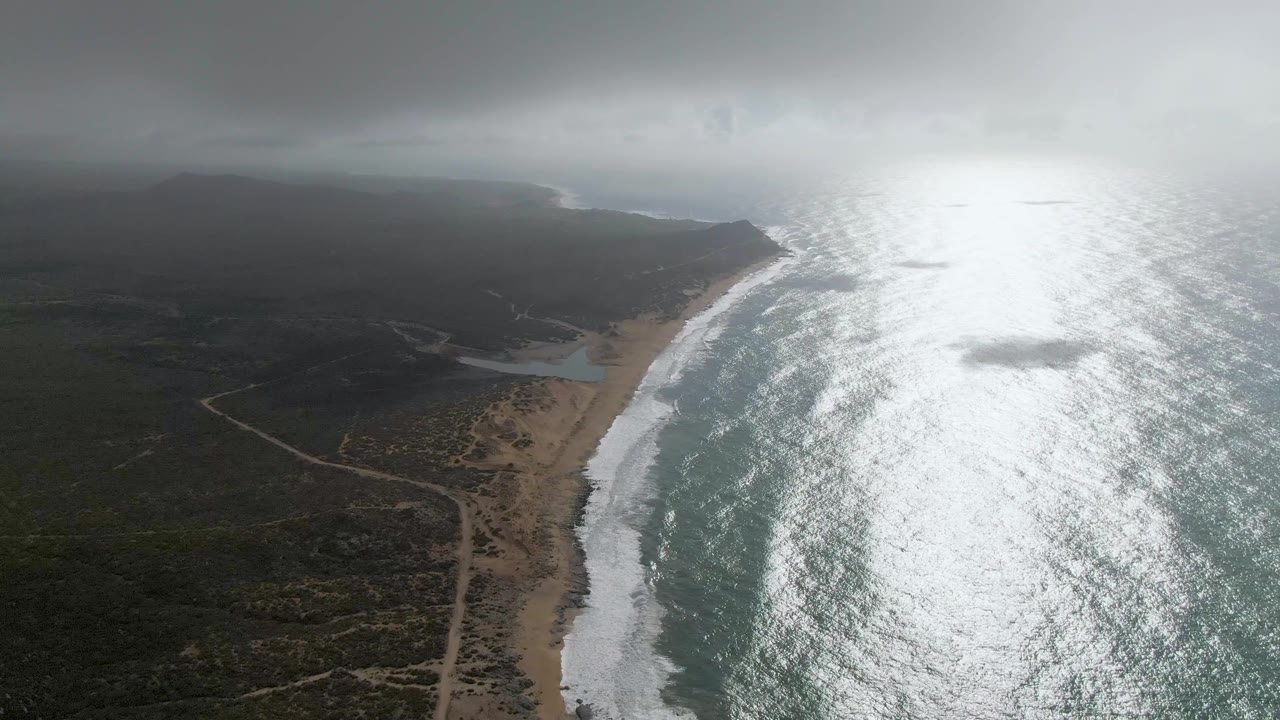 Aerial frame over Punta Gorda East Cape showing the surf line beside dark vegetation, a pale sandy beach, curving dirt roads, intense ocean glare, and no visible people.