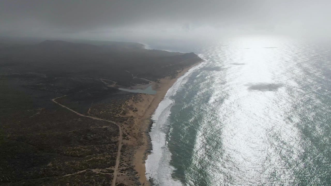 Aerial view along Punta Gorda East Cape with the beach running diagonally through frame, breaking waves to the right, dirt roads over scrubland to the left, clouded horizon, and no visible people.