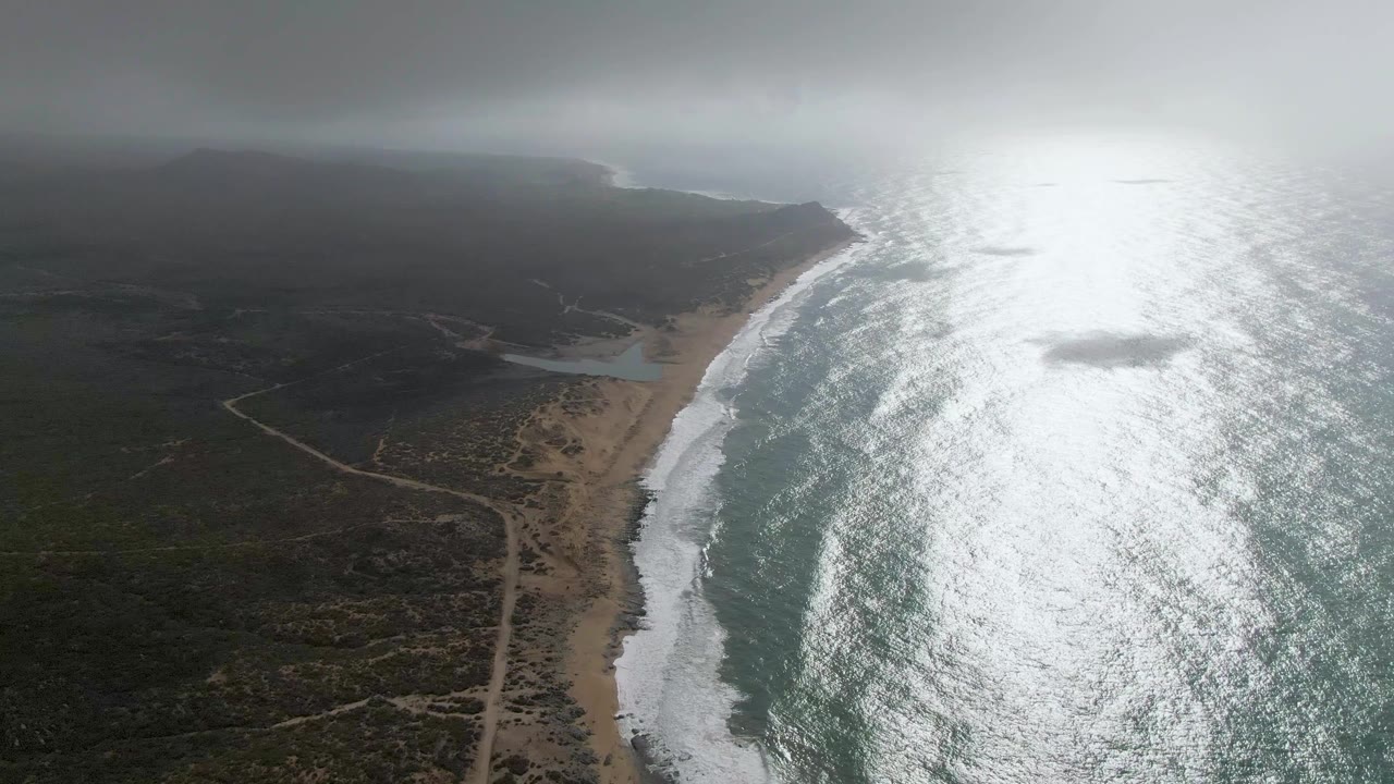 Aerial Punta Gorda East Cape frame with white surf meeting the beach, scrubland and dirt tracks inland, a small inlet or lagoon near the shore, reflective sea, and no visible people.