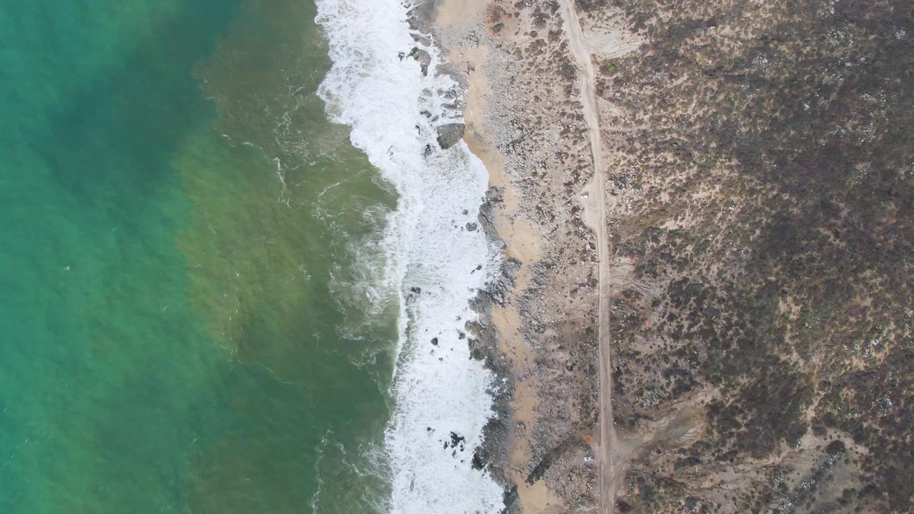 Aerial view in Punta Gorda with green-blue ocean filling the left side, foam sliding onto the sandy rocky edge, dry brushland on the right, a parallel dirt road, and no visible people.