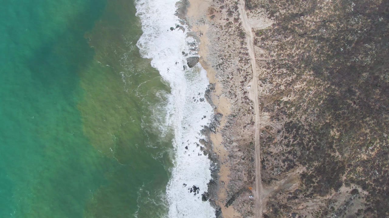 Drone view of turquoise sea, foamy break, sandy rocks, scrubland, and a road along the beach.