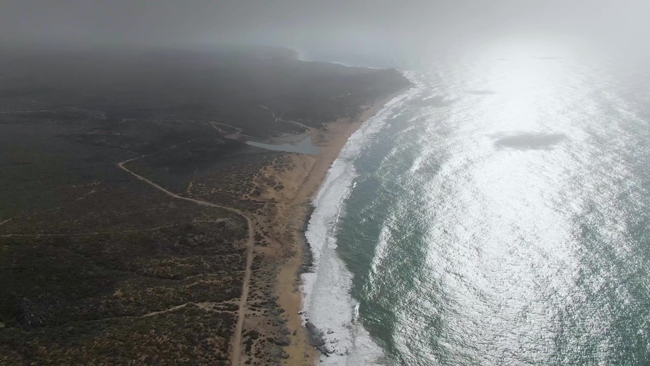 Aerial view over Punta Gorda, Baja California Sur, with white surf along a sandy beach, a dirt road through scrubland, bright ocean glare, mist on the horizon, and no visible people.