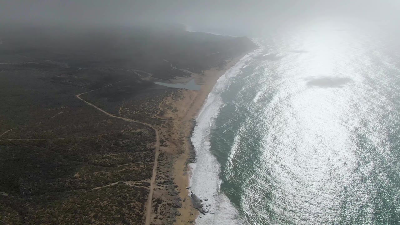 High aerial frame at Punta Gorda showing the foamy shoreline curving past a sandy beach, a pale road crossing dark scrubland, bright water reflection, and no visible people.