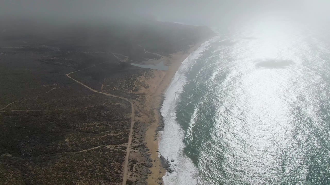 Aerial view over Punta Gorda with the ocean filling the right side, white surf tracing the beach, a narrow road in the inland scrub, heavy glare on the water, and no visible people.