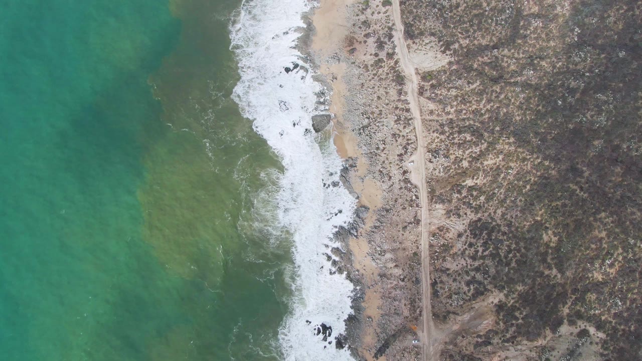 Overhead view of Punta Gorda East Cape coastline with green-blue water, white surf on a sandy rocky edge, a parallel road on the right, and no visible people.
