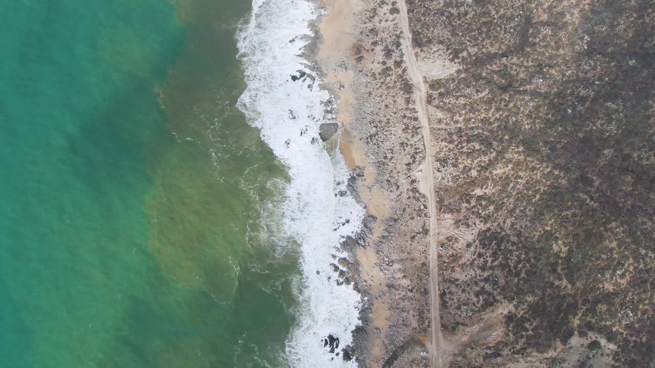 Aerial frame from Punta Gorda East Cape showing surf foam pushed farther along the sandy shore, shallow green water offshore, scrubland inland, and no visible people.