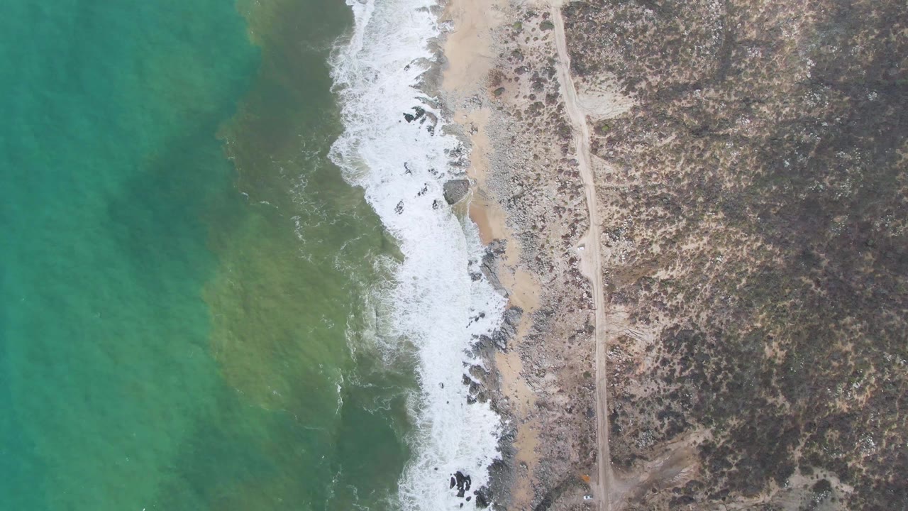 Top-down Punta Gorda East Cape view with a bright band of surf breaking over rocks, sandy patches along the waterline, a straight road inland, and no visible people.