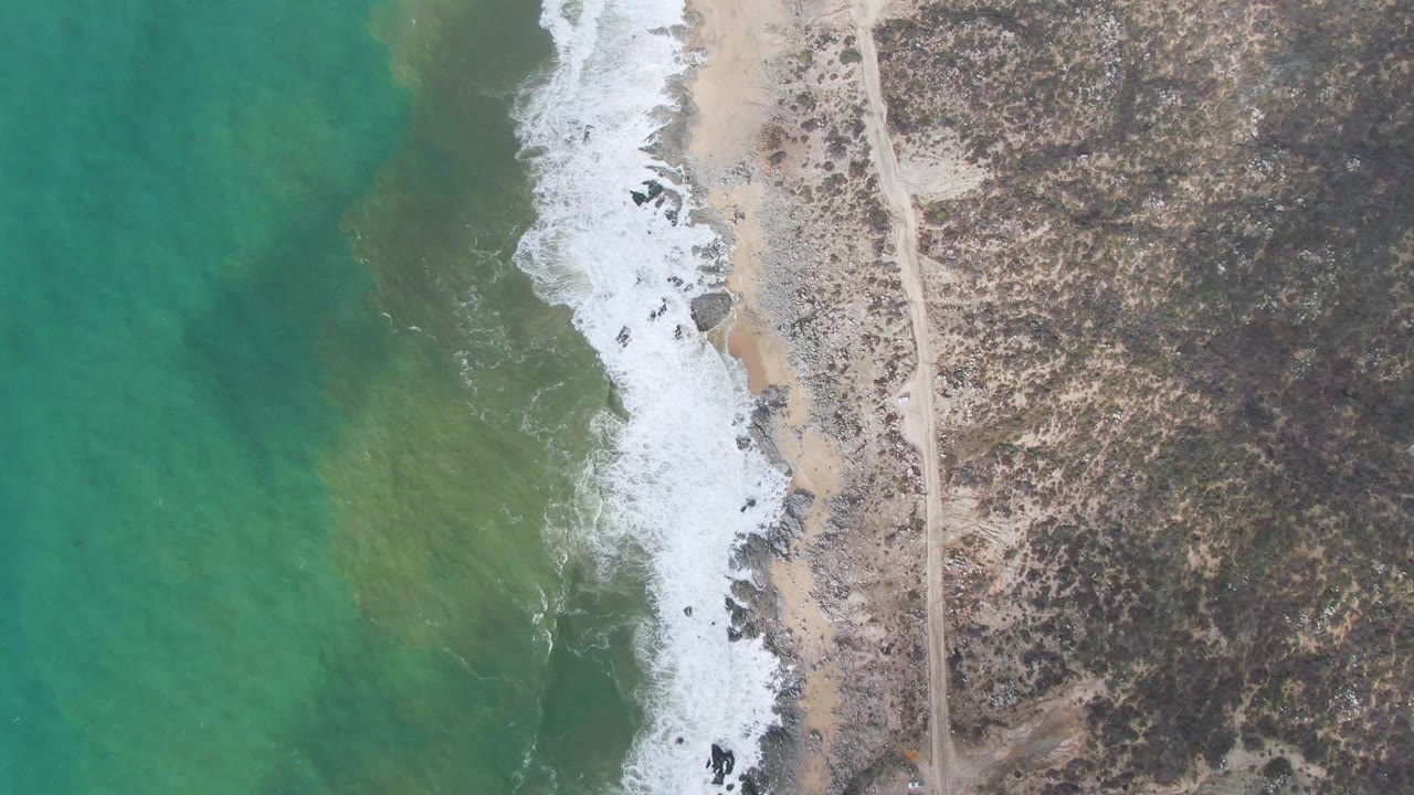 Drone view of turquoise water washing onto sandy rocky shoreline near a road