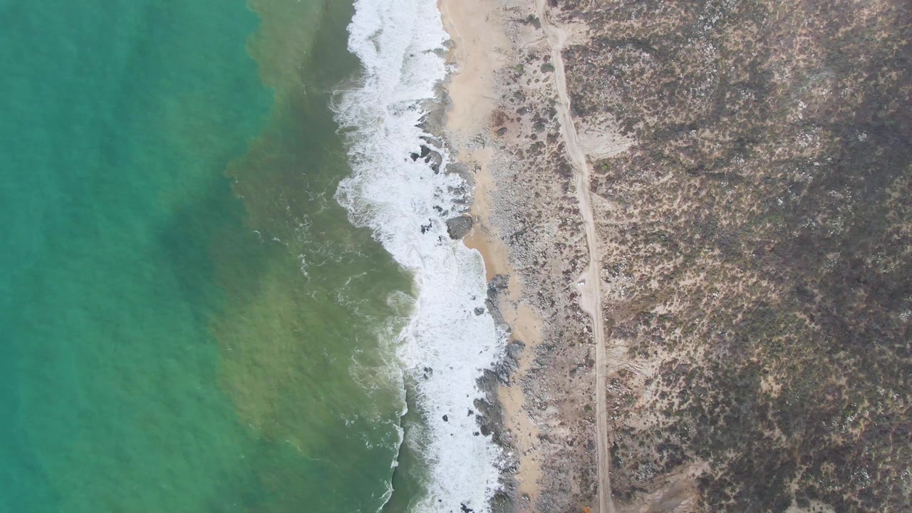 Overhead turquoise water and waves beside a sandy rocky coast road.