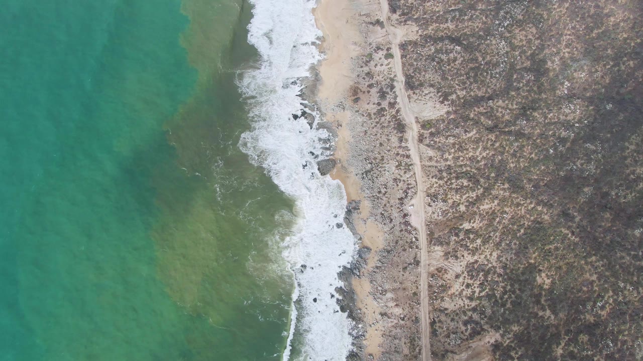 Aerial frame over Punta Gorda coast where a band of white foam pushes against the sandy and rocky shoreline, with green-blue water left and the dirt road still visible inland, no visible people.