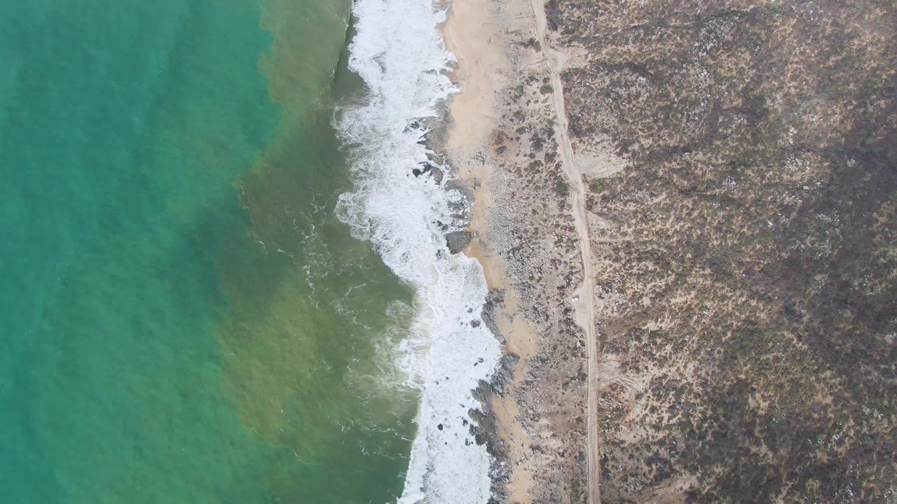Top-down Punta Gorda shoreline frame with brighter turquoise water, broken white surf, tan sand, scattered dark rocks, dry scrubland, and a straight dirt road along the coast, no visible people.