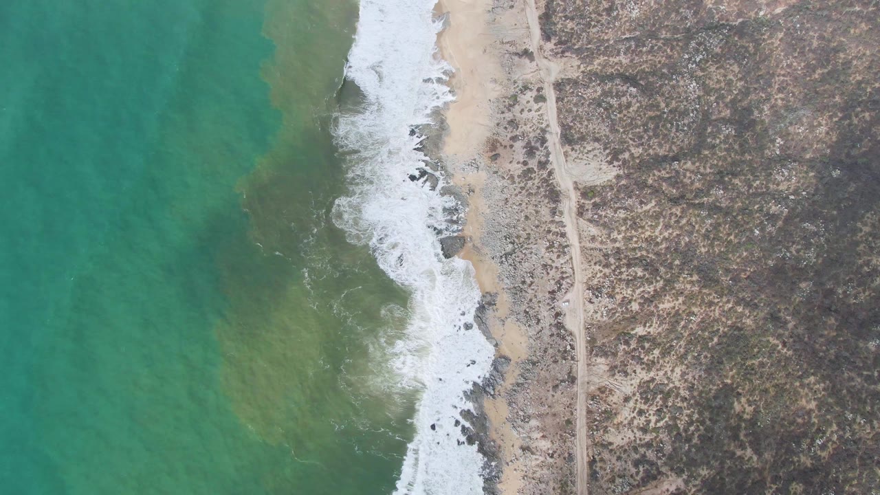 Overhead view in Punta Gorda with more open turquoise water on the left, a foamy wave line through the center, and scrubby rocky land with a faint road on the right, no visible people.