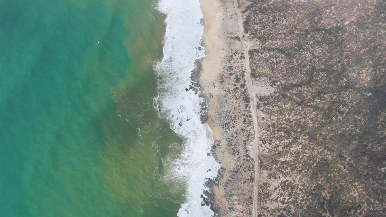 Overhead coastline with turquoise water, white surf, sandy beach, and dry rocky land.