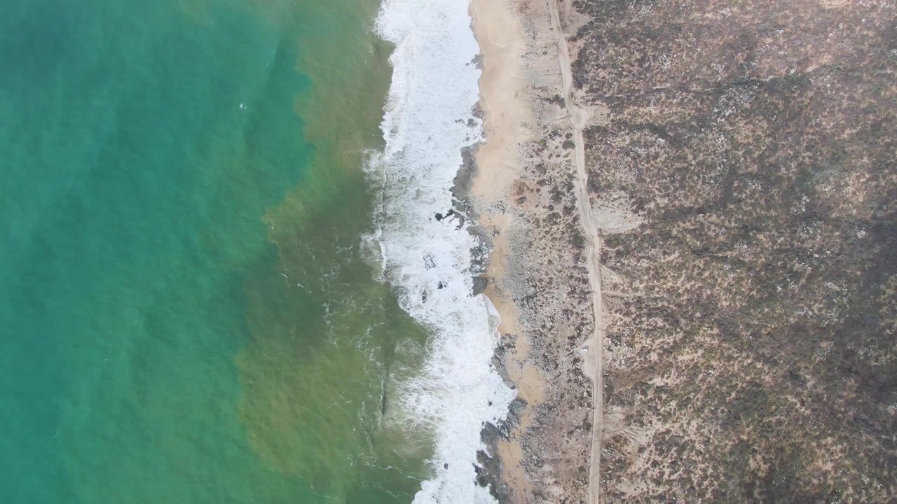 Aerial frame over Punta Gorda East Cape showing white foam advancing along the beach edge, green-turquoise shallows, dry scrubland, and no visible people.