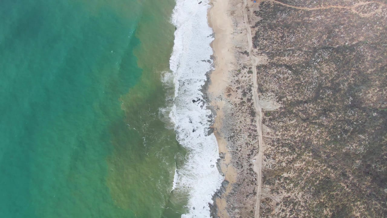 Overhead view at Punta Gorda East Cape with foam spread farther along the sand, turquoise water on the left, dry rocky land on the right, and no visible people.