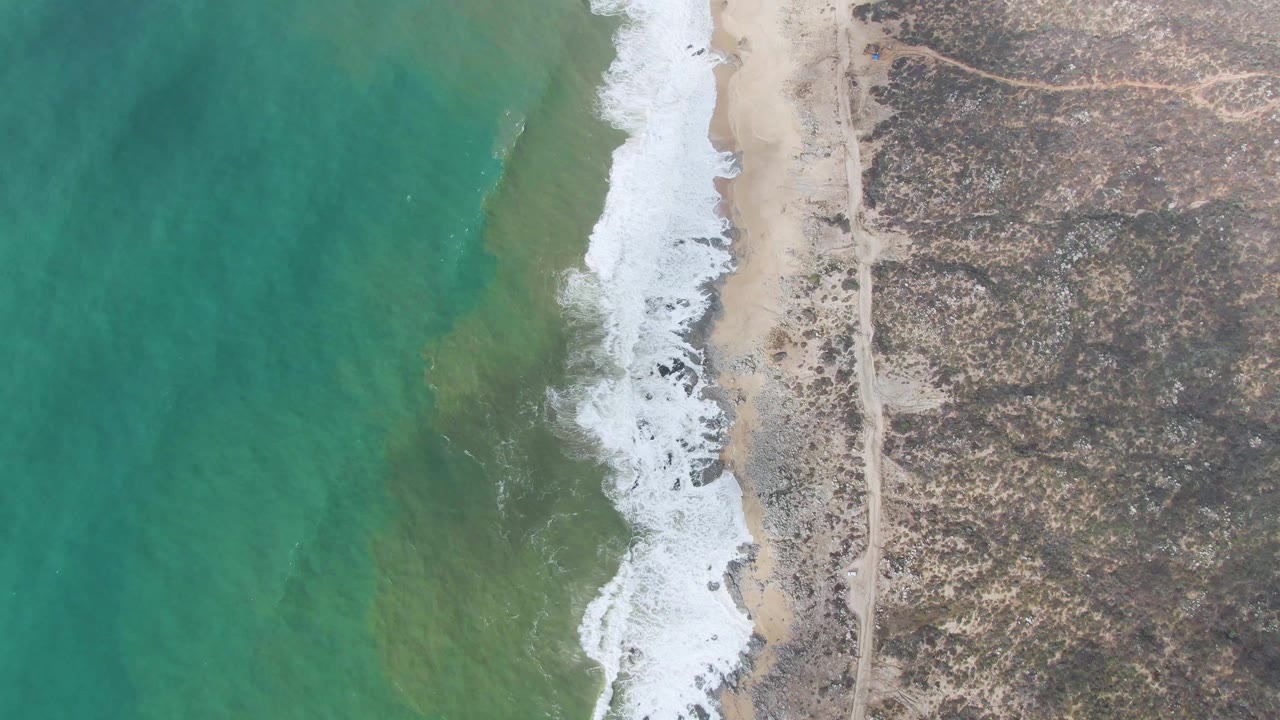 Overhead view at Punta Gorda, East Cape, Baja California Sur, with turquoise water on the left, white surf breaking on sand, rocky terrain on the right, and no visible people.