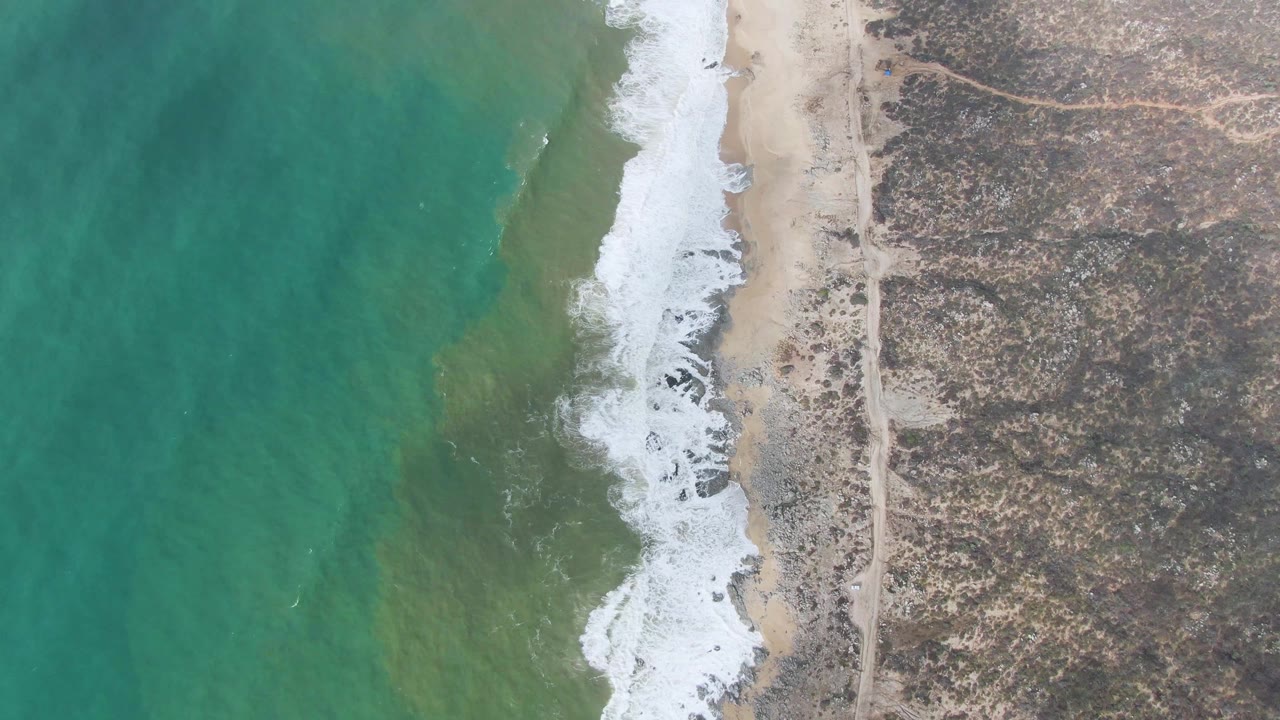 Aerial frame from Punta Gorda, East Cape, Baja California Sur, showing a broad green-turquoise water field, a foamy wave line on the sand, rocky scrubland, and no visible people.
