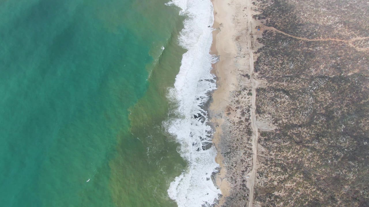 Top-down Punta Gorda shoreline view with white foam curling over a sandy strip, darker rocks near the break, turquoise sea to the left, dry terrain to the right, and no visible people.
