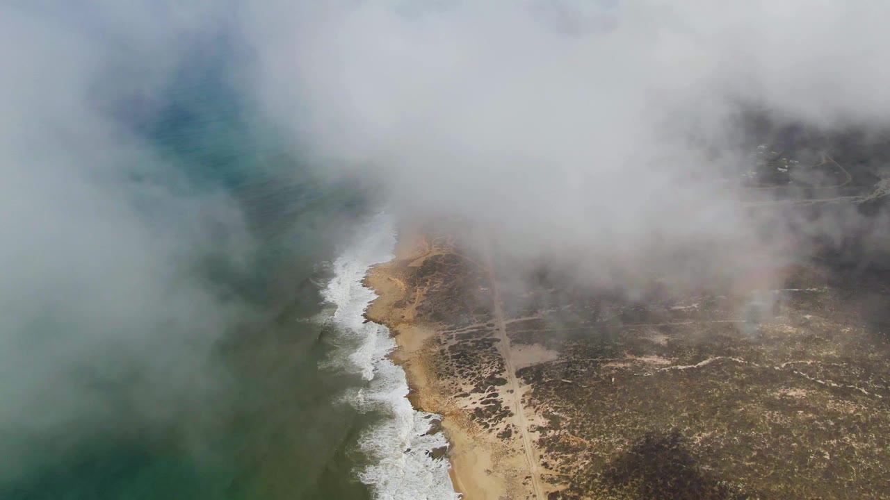 Aerial frame from Punta Gorda shows fog over the ocean and beach, with a bright surf line separating water from dry coastal land and no visible people.