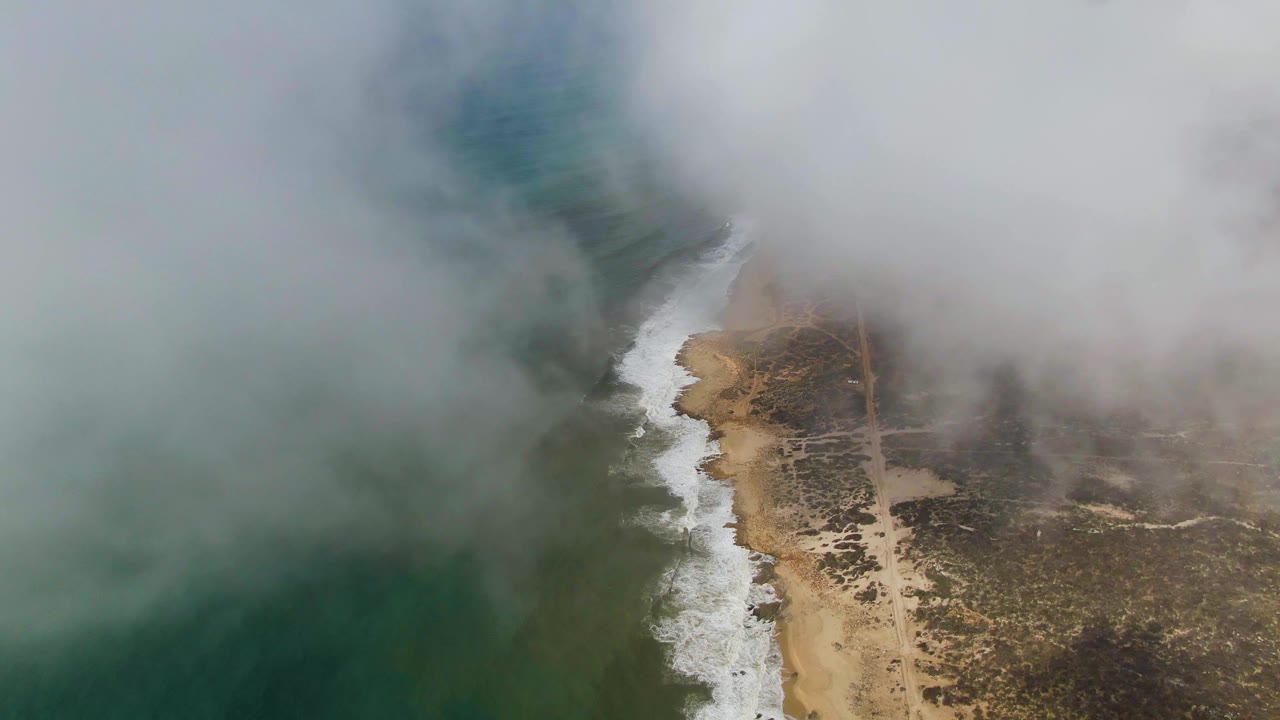 Aerial view at Punta Gorda, Baja California Sur, with fog covering the upper coastline, green ocean on the left, white surf along sand, scrubland on the right, and no visible people.