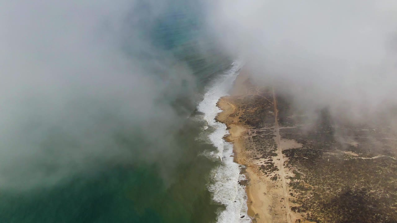 Aerial frame of the Punta Gorda shoreline with thick mist over the top of the image, darker green water at left, a white surf line, sandy beach, inland scrub, and no visible people.