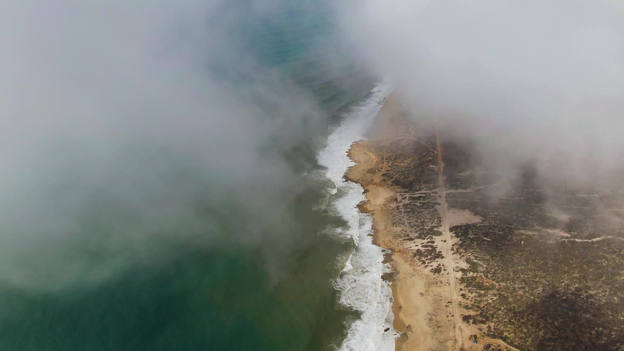 Fog spreads across the upper Punta Gorda coast as the green ocean, bright white wave foam, tan sand, dark scrubland, and a faint path remain visible below, with no visible people.