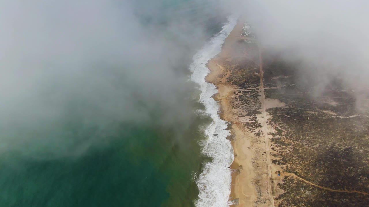 Aerial coastline with fog over ocean, surf, sandy beach, and scrubland.