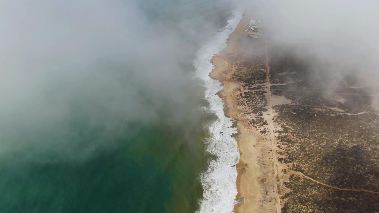 Aerial frame over Punta Gorda East Cape with thicker fog over the ocean, a bright line of breaking waves, tan sand, inland scrub, dirt tracks, and no visible people.