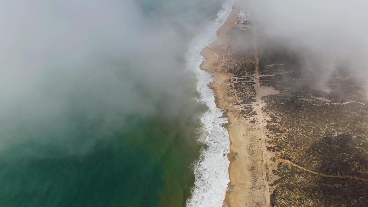 Punta Gorda East Cape appears from above with green water on one side, a continuous strip of white foam, tan beach, brushy inland ground, fog overhead, and no visible people.