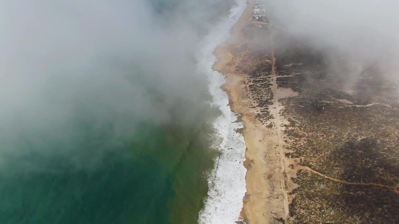Aerial view of Punta Gorda East Cape with fog softening the upper coast, ocean water at left, surf breaking down the beach, inland scrub and faint paths at right, and no visible people.