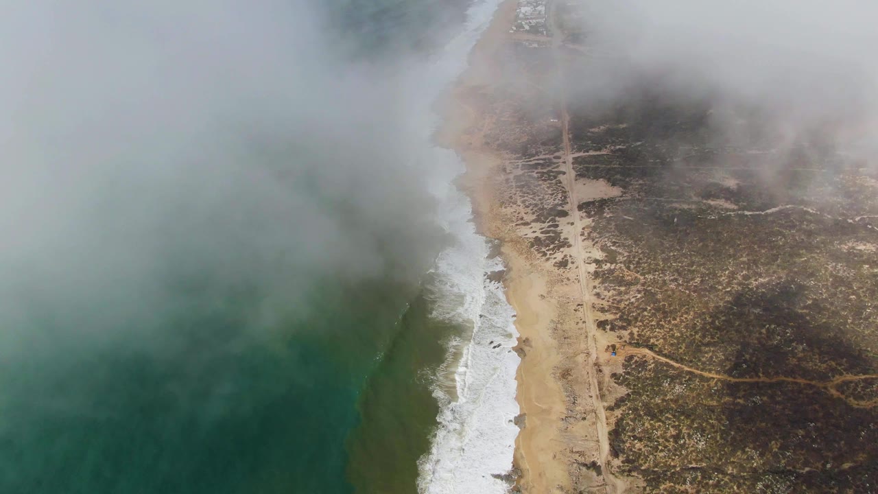 Aerial view of Punta Gorda coastline with green ocean on the left, white surf beside a sandy beach, a straight inland road, low fog overhead, and no visible people.