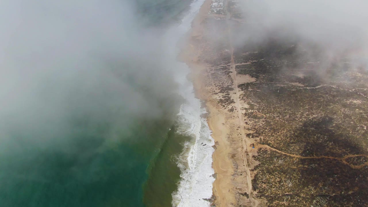 Fog drifts over a sandy shoreline with waves and a parallel road.
