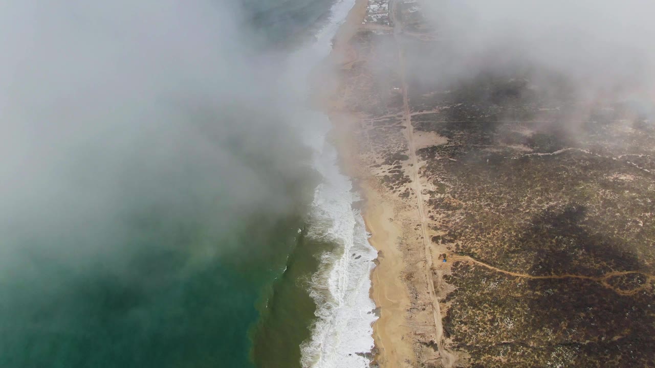 High drone view over Punta Gorda with dense fog crossing the top of frame, darker ocean water to the left, a narrow surf band, sandy beach, road, and no visible people.