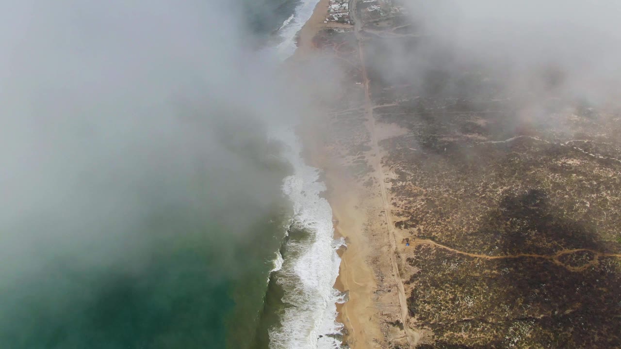 Aerial view over Punta Gordo in Baja California Sur with green-blue ocean on the left, surf along a sandy beach, a coastal road on dry land, low fog overhead, and no visible people.