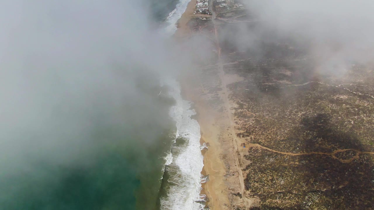 Aerial frame from Punta Gordo showing white surf tracing the beach, a narrow road across scrubby coastal land, fog covering the upper shoreline, and no visible people.