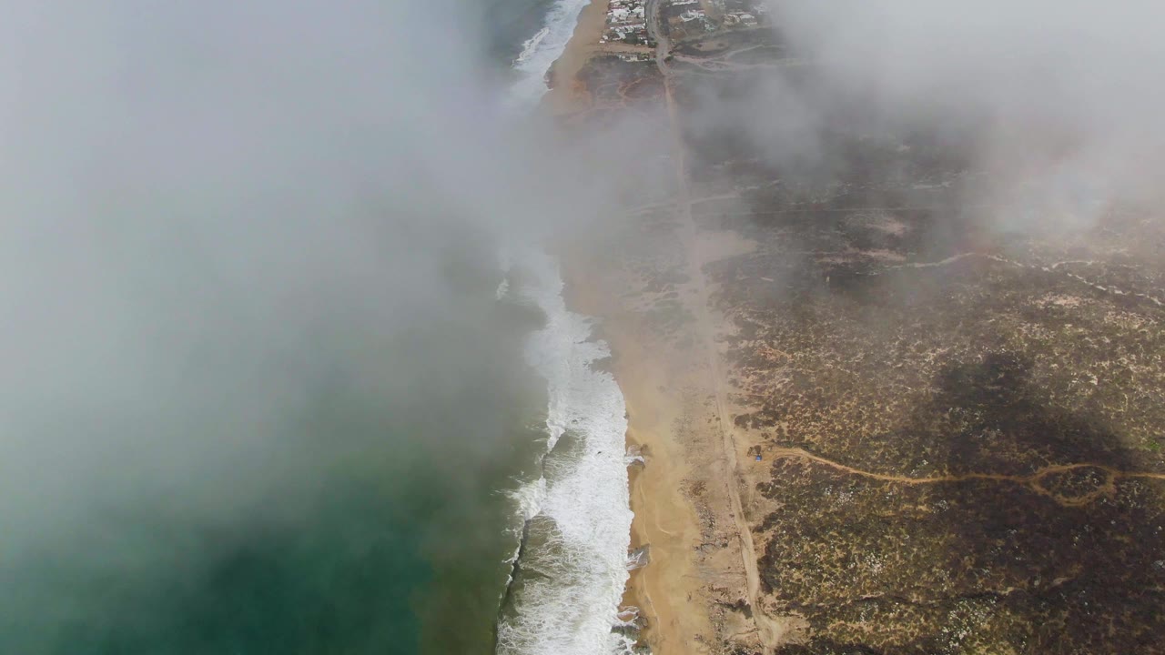 Punta Gordo aerial still with the surf line bright against the beach, the road and brown coastal terrain to the right, thick fog over the upper frame, and no visible people.