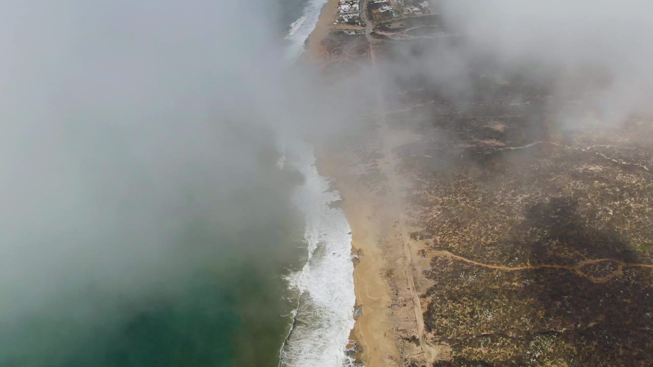 Aerial coastline with heavier fog over ocean, beach, road, and scrubland.