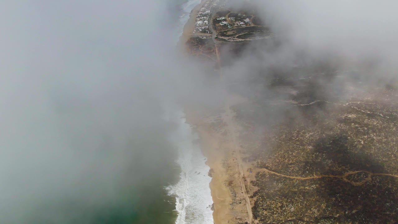 Aerial view of fog drifting over the Punta Gorda coastline, with green-blue ocean at left, a white surf line, sandy beach, brown terrain, and no visible people.