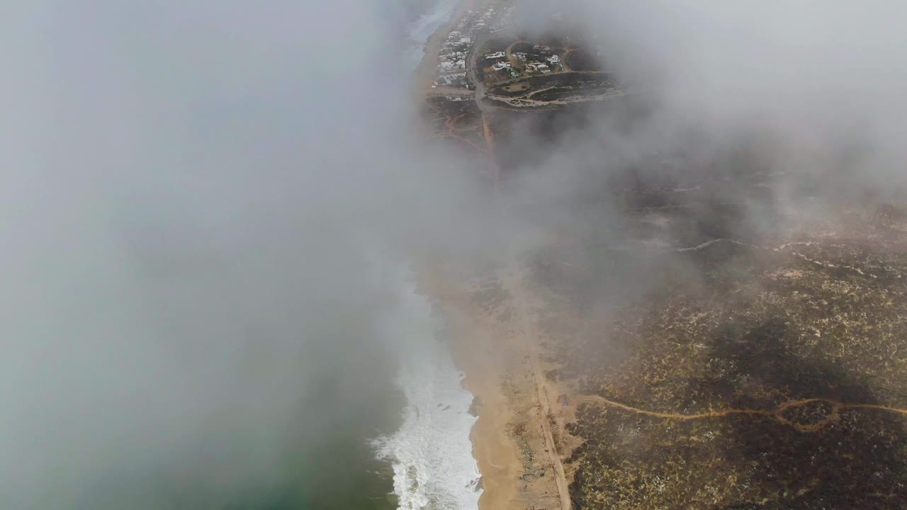 Fog thickens across the Punta Gorda beach from above, leaving the ocean edge and pale surf visible while dry land remains partly exposed, with no visible people.