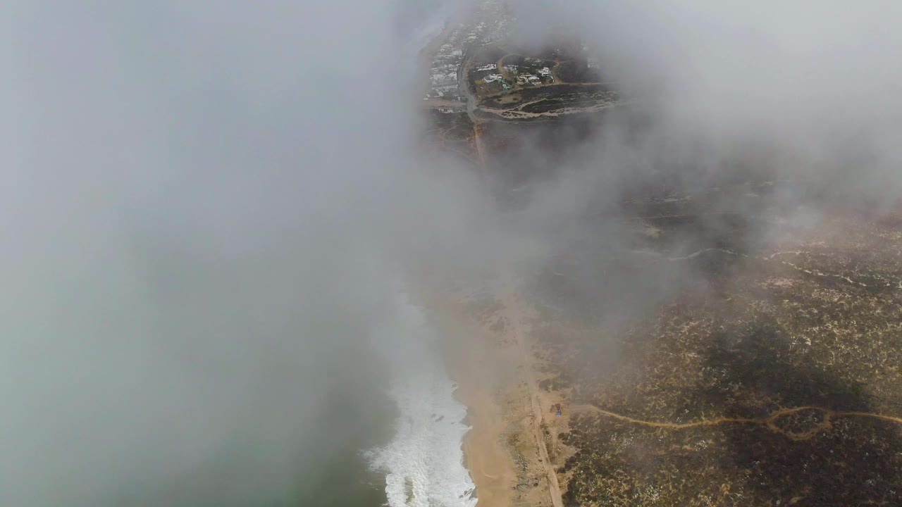 Aerial frame of the Punta Gorda coast with dense mist over the upper frame, muted ocean on the left, a narrow white surf band, exposed beach, and no visible people.